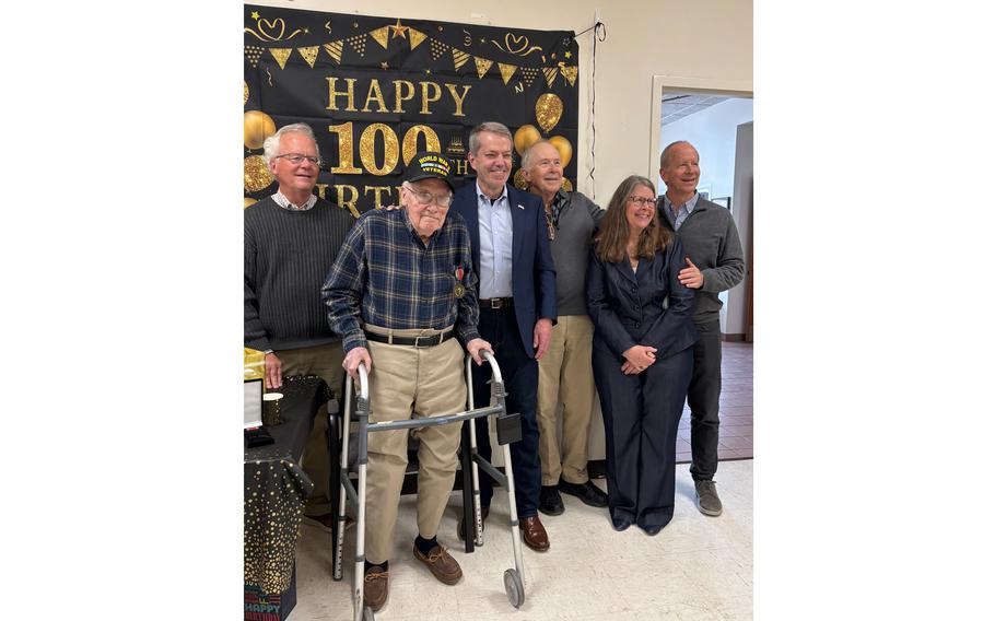Six people standing and smiling for a photo in front of a poster that celebrates a 100th birthday.