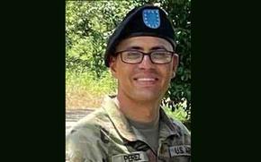 A soldier in camouflage uniform, black beret and black-rimmed glasses smiles while looking forward and posing in front of a tree.