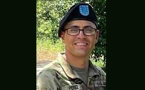 A soldier in camouflage uniform, black beret and black-rimmed glasses smiles while looking forward and posing in front of a tree.