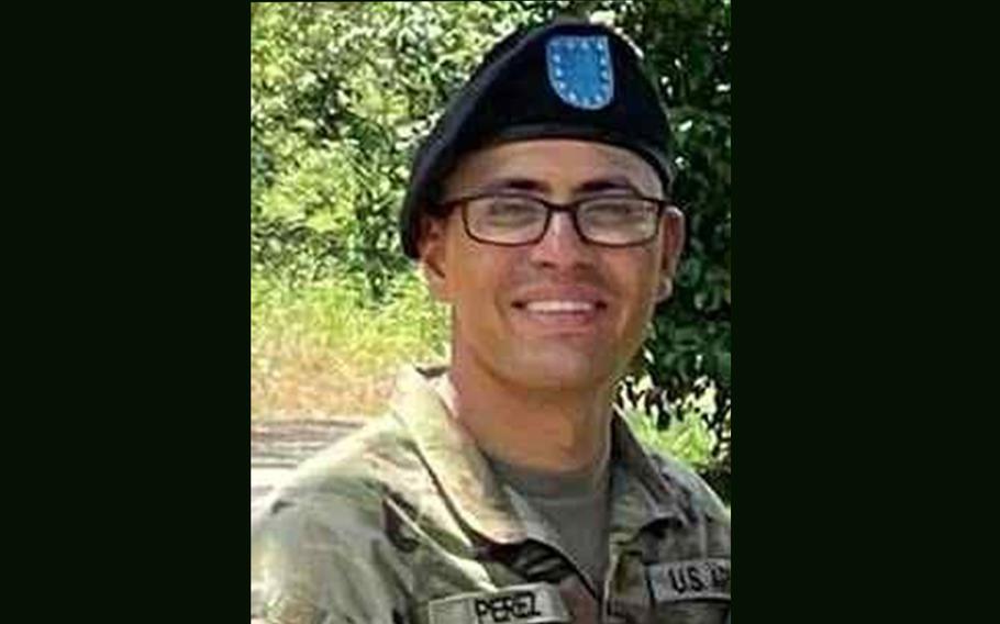 A soldier in camouflage uniform, black beret and black-rimmed glasses smiles while looking forward and posing in front of a tree.