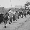 A black-and-white photo of a line of men carrying poles that hold wrapped corpses. 