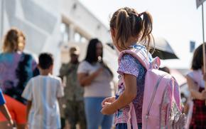 A girl wearing pigtails and wearing a pink backback looks around a school yard.