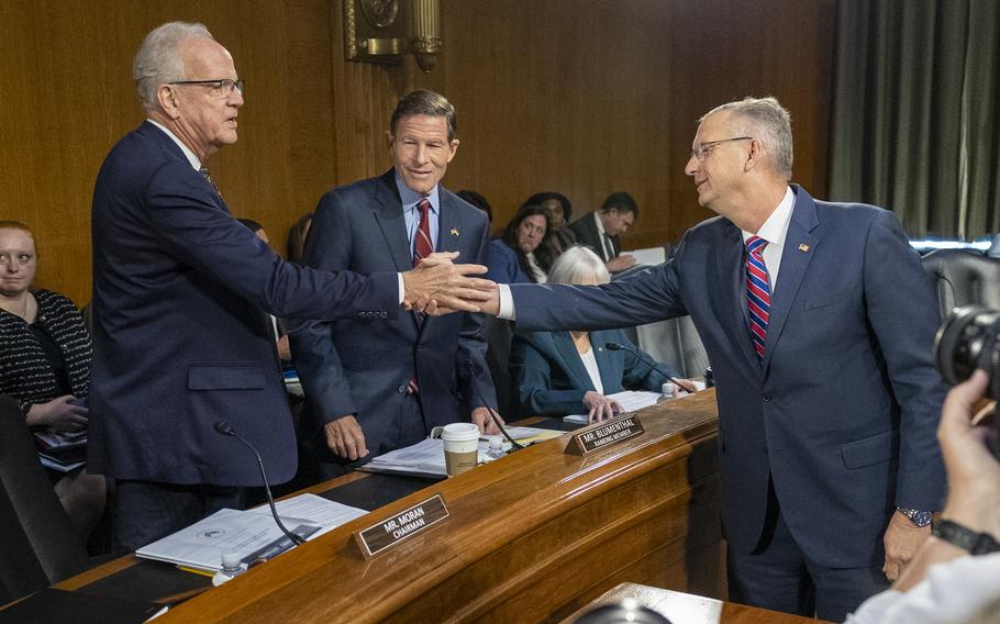 Two men in suits shake hands in a congressional hearing room as photographers take pictures and a lawmaker looks on.