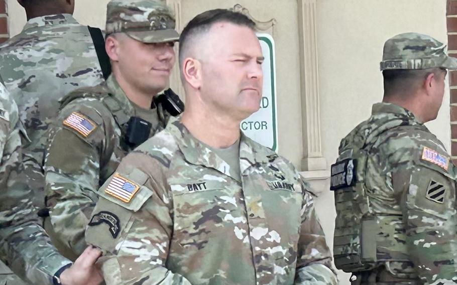 An Army soldier in camouflage uniform stands with a hat in his hands in front of building, with other soldiers in camouflage behind him.