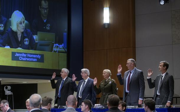 Five people stand with their hands raised as a monitor overhead displays a woman’s face.