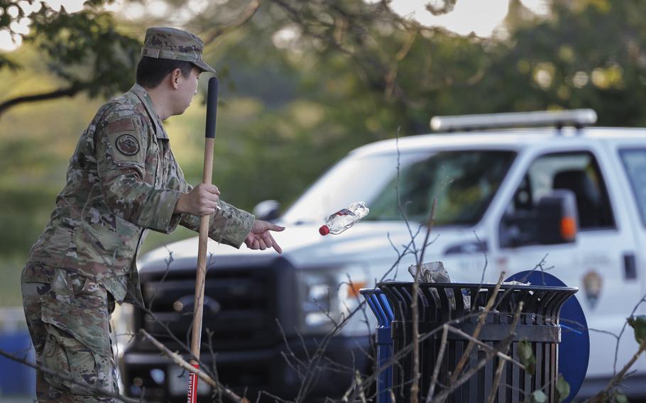 A soldier tosses a bottle into a trashcan with one hand; the other hand holds something for cleaning.
