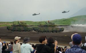 Japan Ground Self-Defense Force tanks perform live-fire drills in front of spectators while helicopters fly in the background.