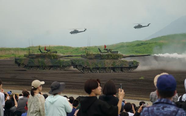 Japan Ground Self-Defense Force tanks perform live-fire drills in front of spectators while helicopters fly in the background.