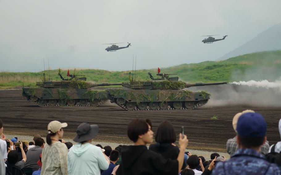 Japan Ground Self-Defense Force tanks perform live-fire drills in front of spectators while helicopters fly in the background.