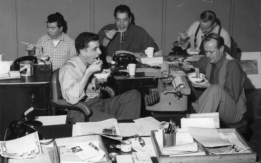 A black and white photo of five men eating ramen at their desks.