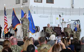 A uniformed officer stands at attention and salutes in front of four flags, including the U.S. flag and the Guam territorial flag. Three other military officers, also in uniform, are seated on a stage with leather chairs, smiling and observing the proceedings.
