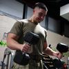 A military member in camouflage pants and olive green shirt lifts dumbbells with each hand while exercising in a gym.