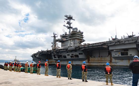 Sailors wearing hard hats and red lifeguard vests over their uniforms stand in a row facing the carrier.