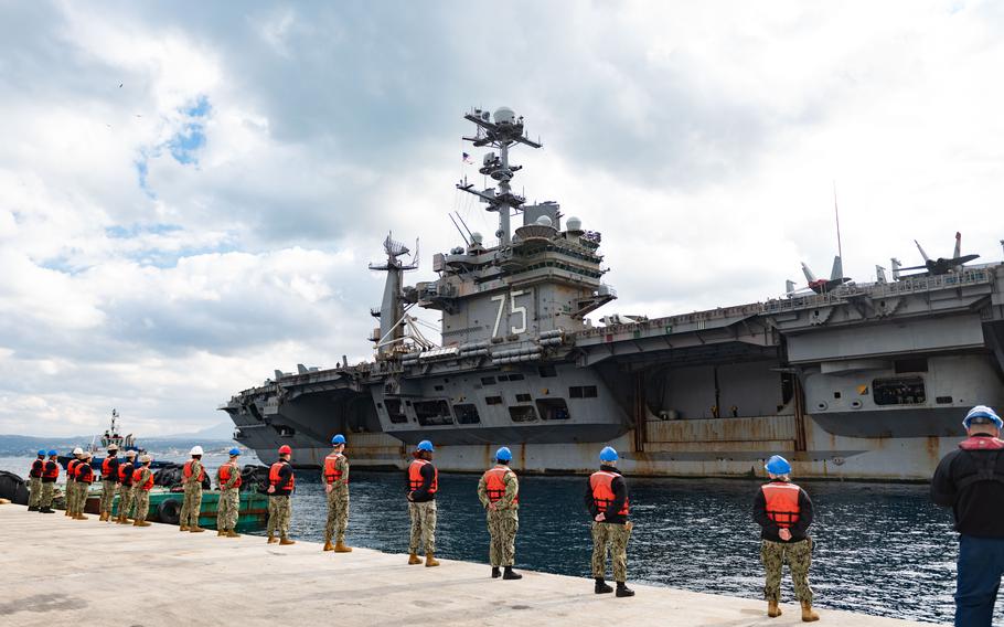 Sailors wearing hard hats and red lifeguard vests over their uniforms stand in a row facing the carrier.