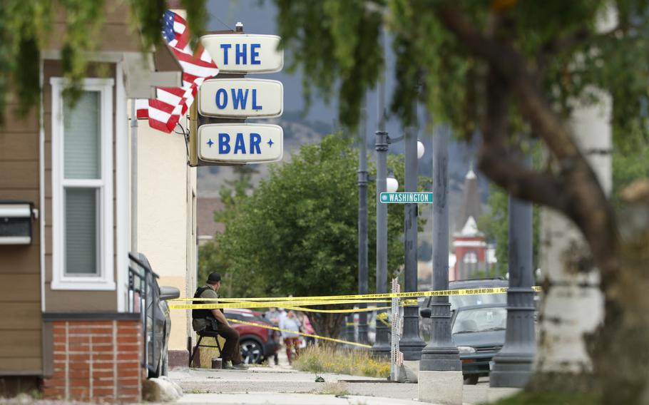Yellow police crime scene tape is wrapped around lightposts on a sidewalk next to a building with a white sign reading “The Owl Bar” and a U.S. flag.