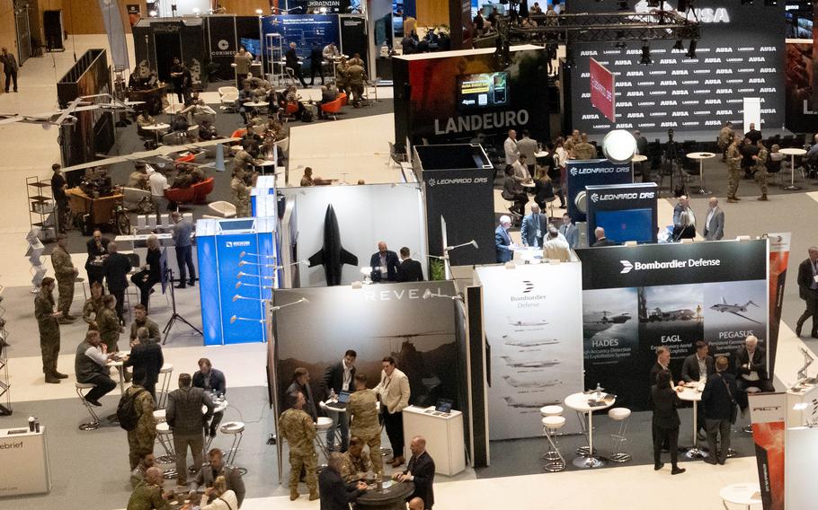 An overhead view of a defense industry exhibition featuring numerous vendor booths and displays from companies like Bombardier Defense with a mix of uniformed service members and industry representatives mingling throughout the venue. 