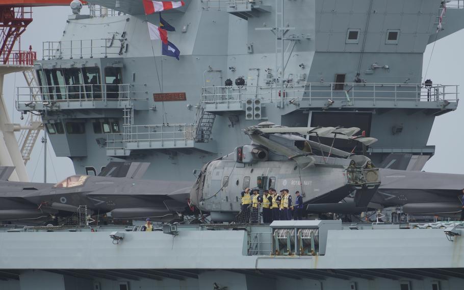 A Royal Navy helicopter is shown on the flight deck of an aircraft carrier, with several crew members in high-visibility yellow jackets gathered around the aircraft. The ship’s superstructure and bridge are visible in the background, along with an F-35 fighter jet parked on the deck.