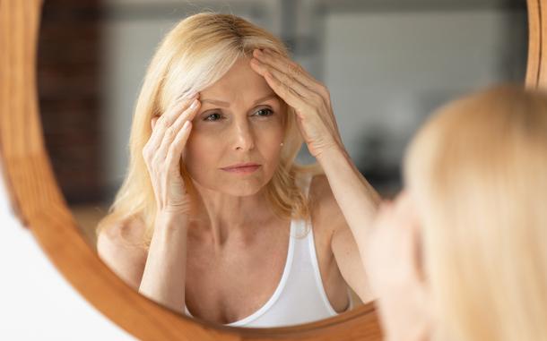 Senior woman looking at mirror and touching forehead, upset aged lady examining fine lines on her face, suffering skin aging, selective focus on reflection