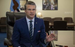 Pete Hegseth sits in an office and speaks while holding a folder in his right hand and gesturing with his left hand. 