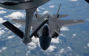 A military fighter jet, seen from the back of a tanker aircraft, attaches a tether for midair refueling with clouds behind and the ground in the distance below.