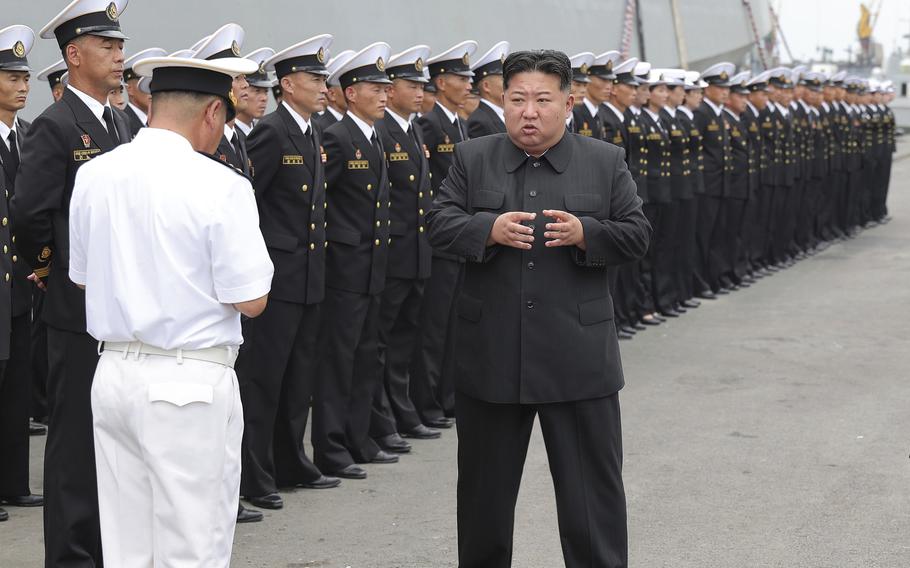 Kim Jong Un, wearing black shirt and pants, stands on the deck of a military warship facing a North Korean naval officer in white dress uniform and in front of a line of North Korean sailors in black dress uniforms standing at attention.