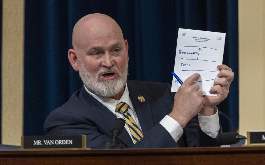 A bearded congressman sits behind a dais holding up a sheet of paper and pointing at notes written on it.
