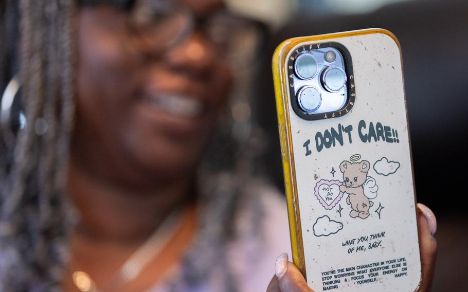 An out-of-focus woman in the background holds up the back of an in-focus iPhone.