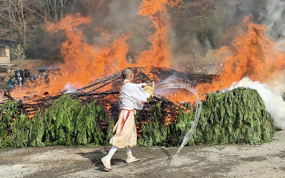 A Japanese monk in traditional attire throws a bucket of water on a large pile of burning tree branches.