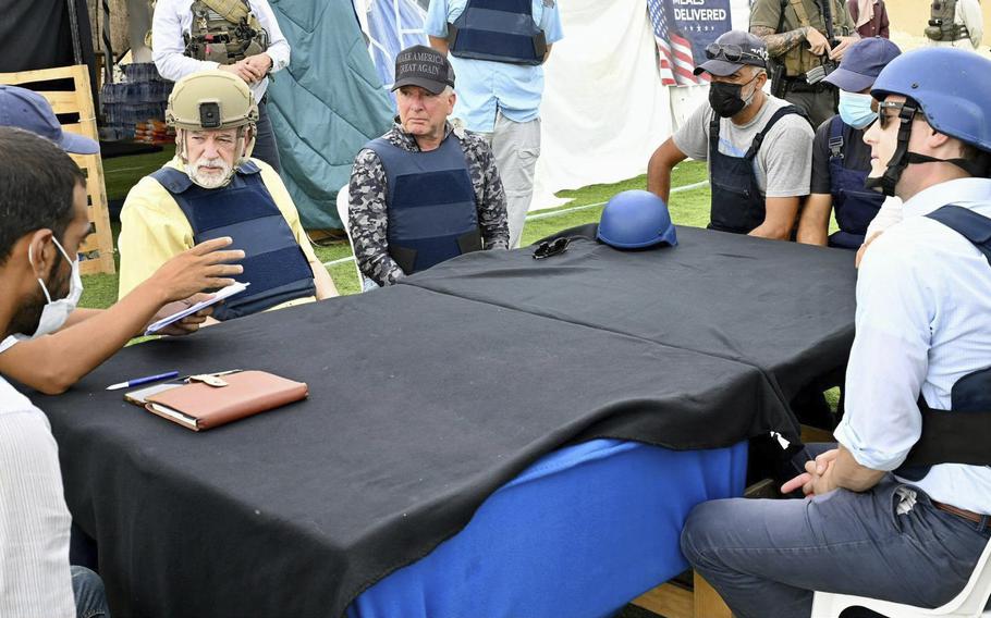 A group of men wearing bulletproof vests and helmets and hats sit around a table covered by a black cloth, with a large multi-colored tent in the background.