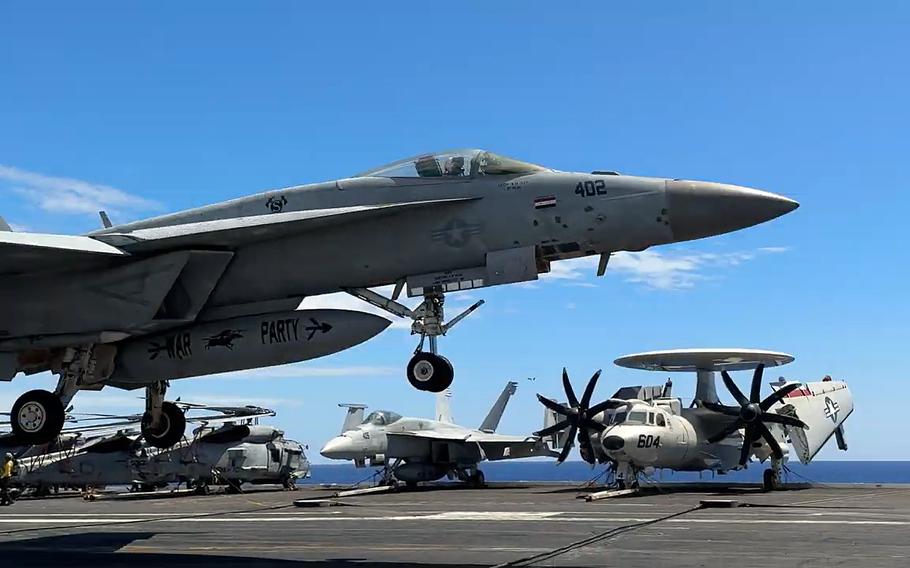 A fighter jet lands on an aircraft carrier with other aircraft visible behind it.