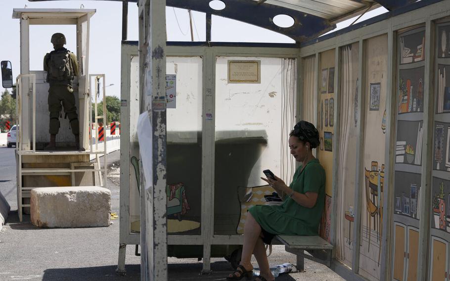 An Israeli soldier stands guard at a bus stop as a woman waits for a bus at the Gush Etzion junction, the transportation hub for a number of West Bank Jewish settlements, Thursday, June 9, 2022. Israeli forces shot and killed a Palestinian man on Sunday, the Palestinian Health Ministry said, after the Israeli military said he tried to illegally cross the separation barrier that divides Israel and the occupied West Bank.