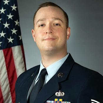 A portrait of an Air Force officer in blue dress uniform posing in front of a U.S. flag.