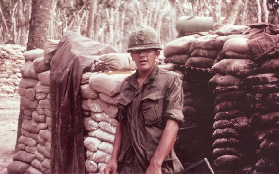A 1960s-era image of a soldier in combat gear standing in front of a stack of sandbags.