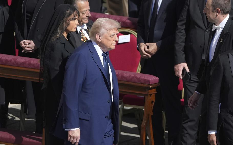 “A man in a blue suit walks past red upholstered chairs at an outdoor formal event, accompanied by a woman dressed in black with a lace veil and another man wearing a medal and smiling. Other attendees in dark suits stand nearby.