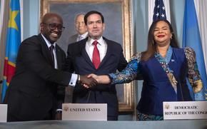 A man in a dark suit shakes hands with a woman in a blue dress, while Marco Rubio stands between them.