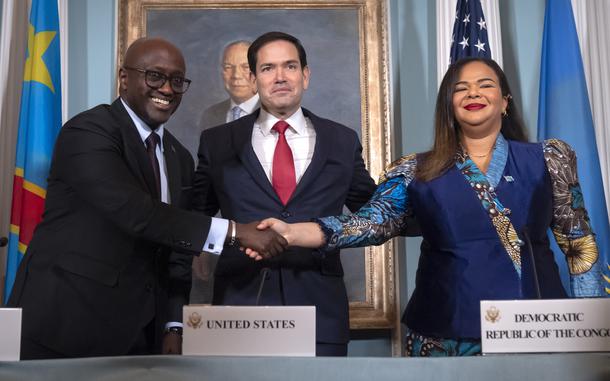 A man in a dark suit shakes hands with a woman in a blue dress, while Marco Rubio stands between them.
