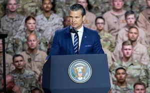 Pete Hegseth stands at a podium with the U.S. seal on it and speaks into a microphone, with military service member seated behind him.