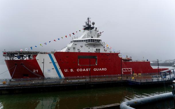 A military ship with a red hull and white lettering reading “U.S. Coast Guard” sits in the water moored to a pier with a foggy sky in the background.