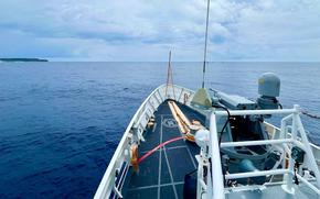 The bow deck of a naval vessel at sea, showing the ship’s forward section with white railings and naval equipment. The vessel is surrounded by deep blue ocean water extending to the horizon, with a distant landmass visible on the left side under a cloudy sky.
