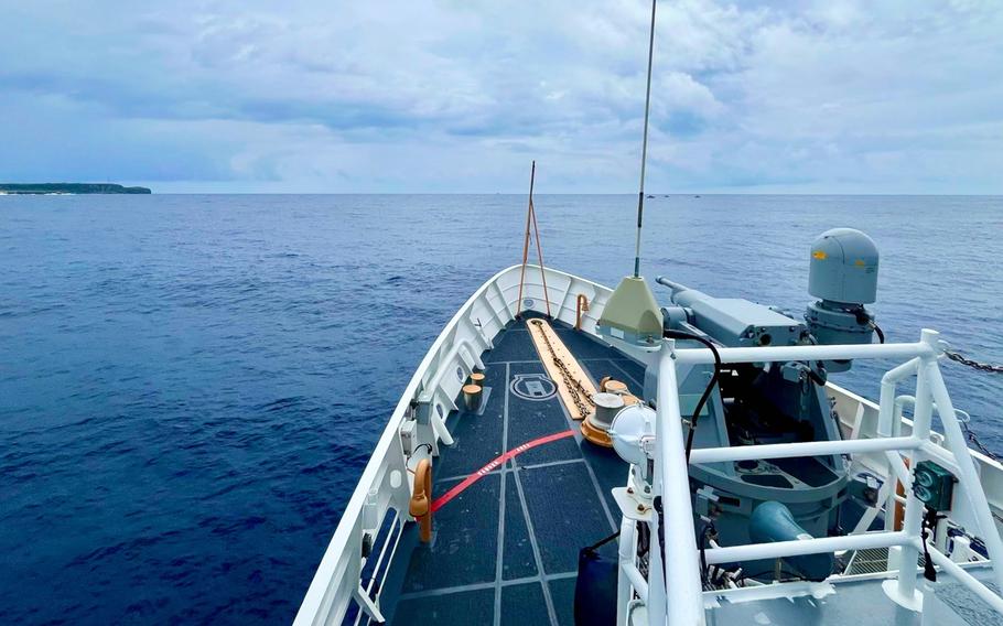 The bow deck of a naval vessel at sea, showing the ship’s forward section with white railings and naval equipment. The vessel is surrounded by deep blue ocean water extending to the horizon, with a distant land mass visible on the left side under a cloudy sky.