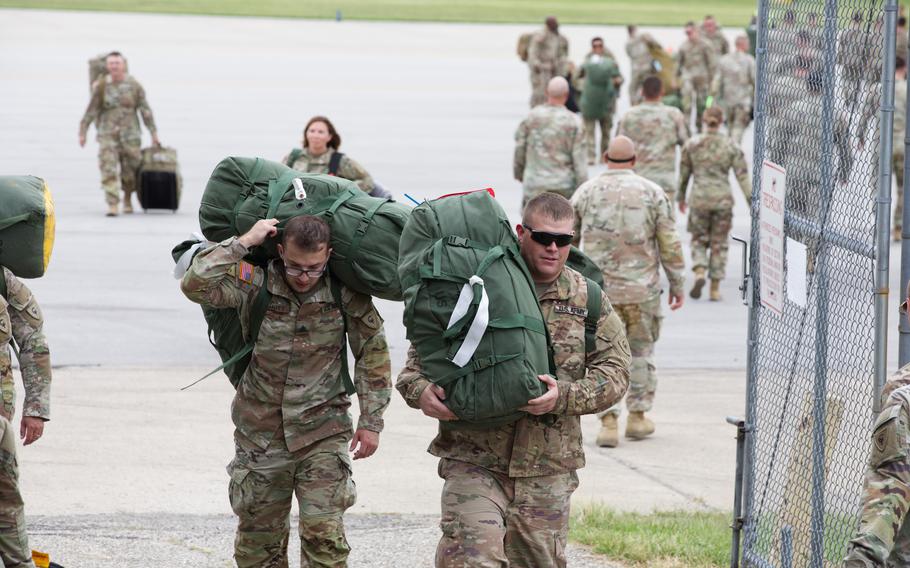 Two soldiers carrying duffel bags in the foreground with other soldiers out-of-focus and milling about behind them.
