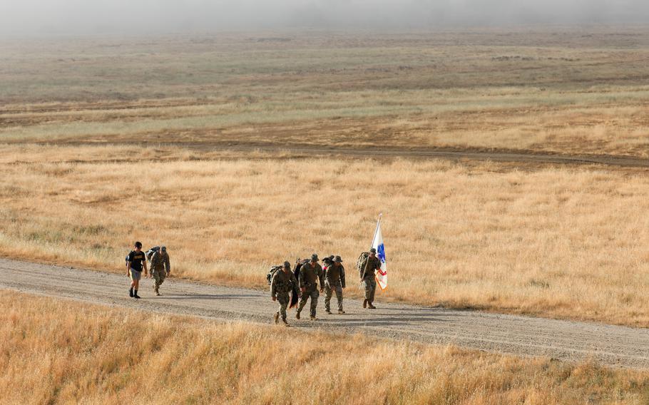 Soldiers and a civilian, seen from a distance, march along a dirt path in a grassy field that stretches into the distance.