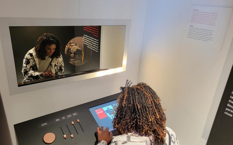 A museum visitor sits in front of a display with a camera and deck with screens and controls. 