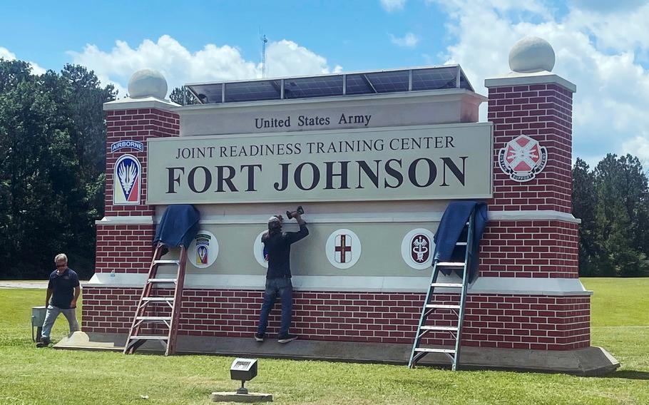 Workers on ladders update the entrance sign to “United States Army Joint Readiness Training Center Fort Johnson,” a brick and concrete structure with military unit emblems and solar panels on top, set against a blue sky with white clouds.