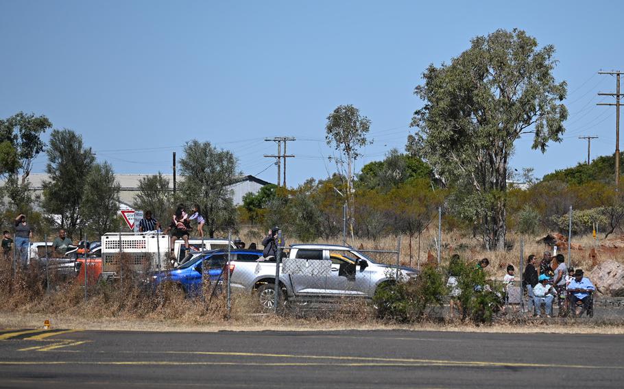 A crowd of people gathered around several pickup trucks and utility vehicles in a rural setting with trees, power lines and dry grassland. 
