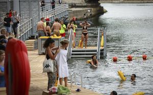 People in swimwear line up on a wooden deck with railings and stairs leading into the river’s water, where several people are swimming among orange buoys. 