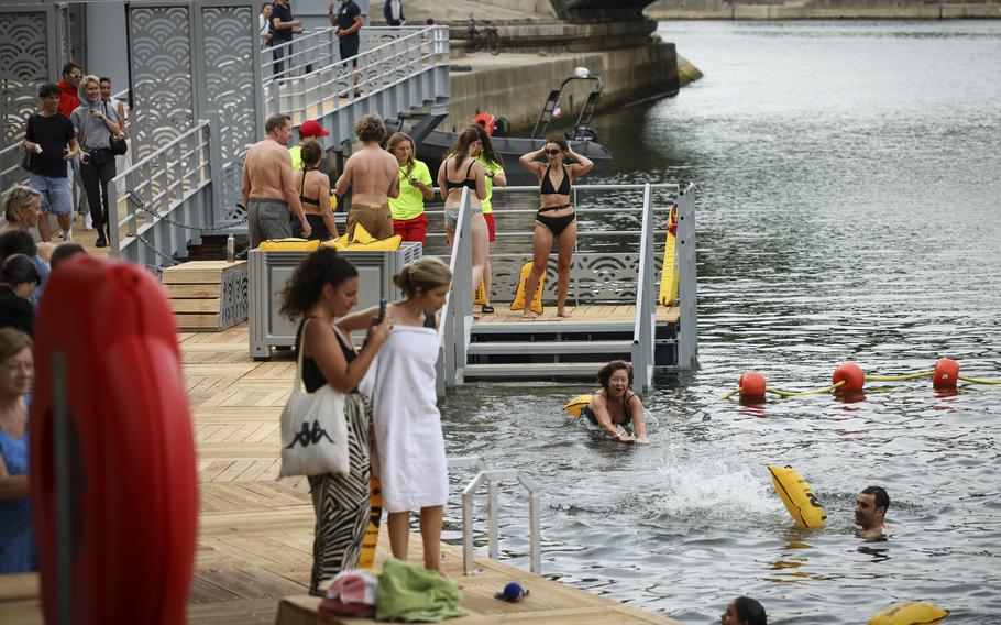 People in swimwear line up on a wooden deck with railings and stairs leading into the river’s water, where several people are swimming among orange buoys. 