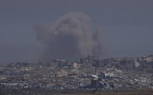 A plume of smoke, seen from a distance, rises into the sky above a landscape of destroyed buildings.