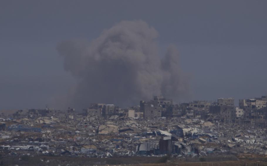 A plume of smoke, seen from a distance, rises into the sky above a landscape of destroyed buildings.