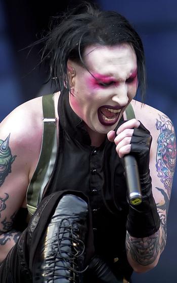 A vertical photo of Marilyn Manson singing or shouting into a microphone that he is holding with one hand.